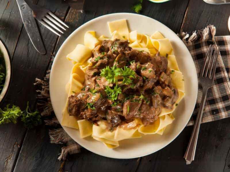 Warm and flavorsome beef stroganoff over cooked egg noodles nicely served on a table with a knife and forks.